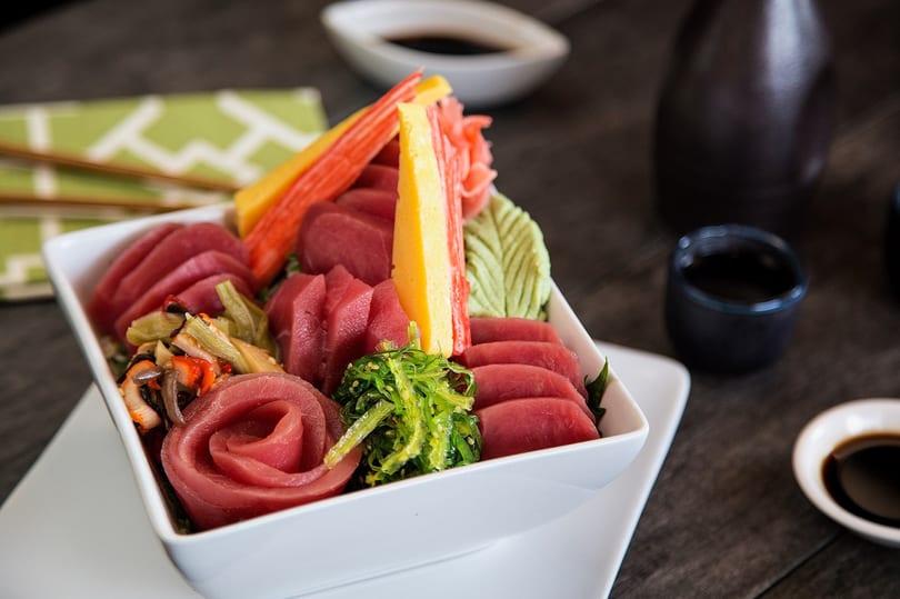 A white bowl filled with various sashimi, including tuna, crab sticks, seaweed salad, and pickled ginger.