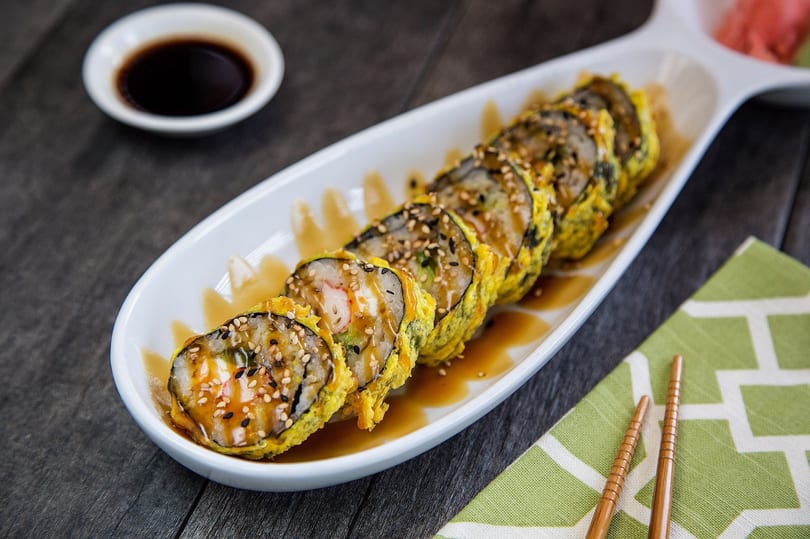 Tempura sushi rolls with sauce and sesame seeds on a white platter, with soy sauce and chopsticks.