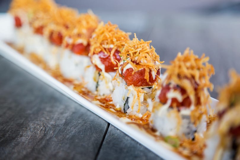 A long white plate of sushi rolls topped with red tuna, white sauce, and crispy fried onions.