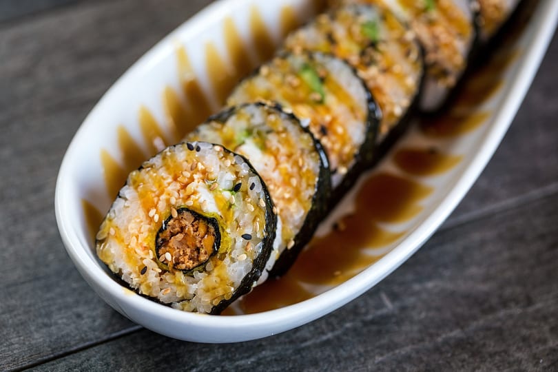 Close-up of sushi rolls with sesame seeds and sauce on a white rectangular plate.