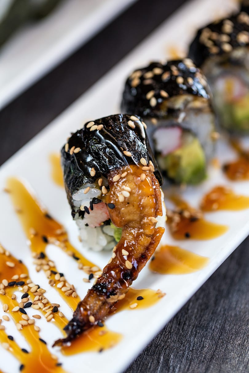 Sushi rolls with eel, avocado, and crab, drizzled with sauce and sesame seeds on a white plate.