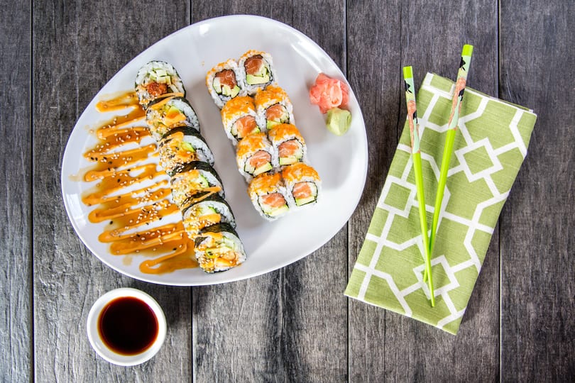 Two rows of sushi rolls with sauces, ginger, wasabi, soy sauce, and chopsticks on a wooden table.
