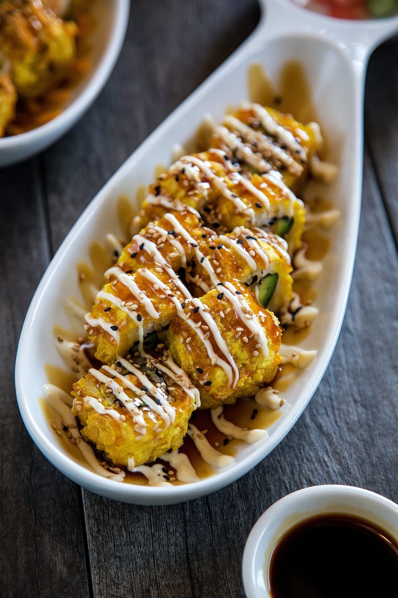 Golden fried sushi rolls drizzled with white and brown sauces, topped with sesame seeds on a white platter.