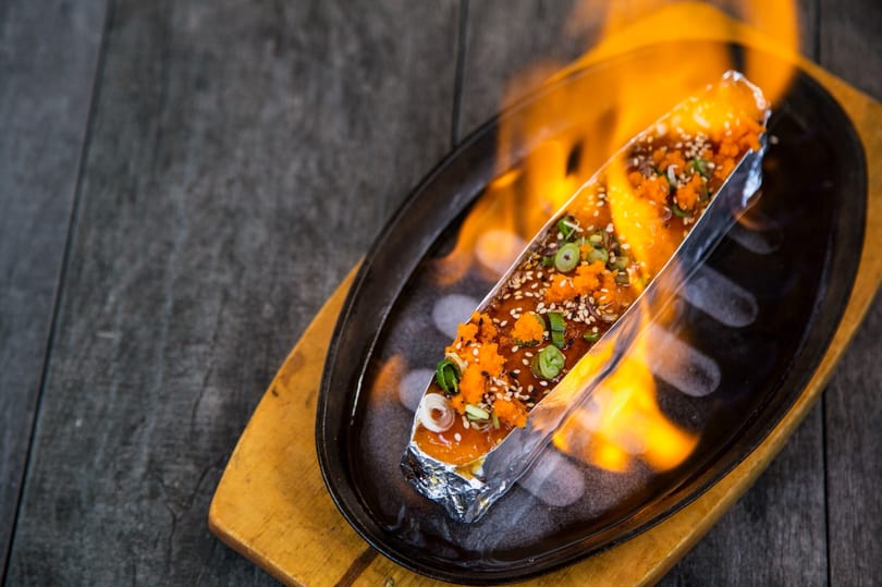 Flaming sushi roll in foil, topped with roe, sesame seeds, and green onions on a hot plate.