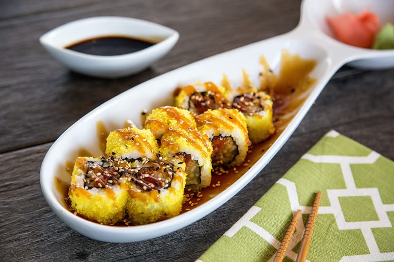 Sushi rolls with yellow topping and sauce on a white plate, with soy sauce and chopsticks.