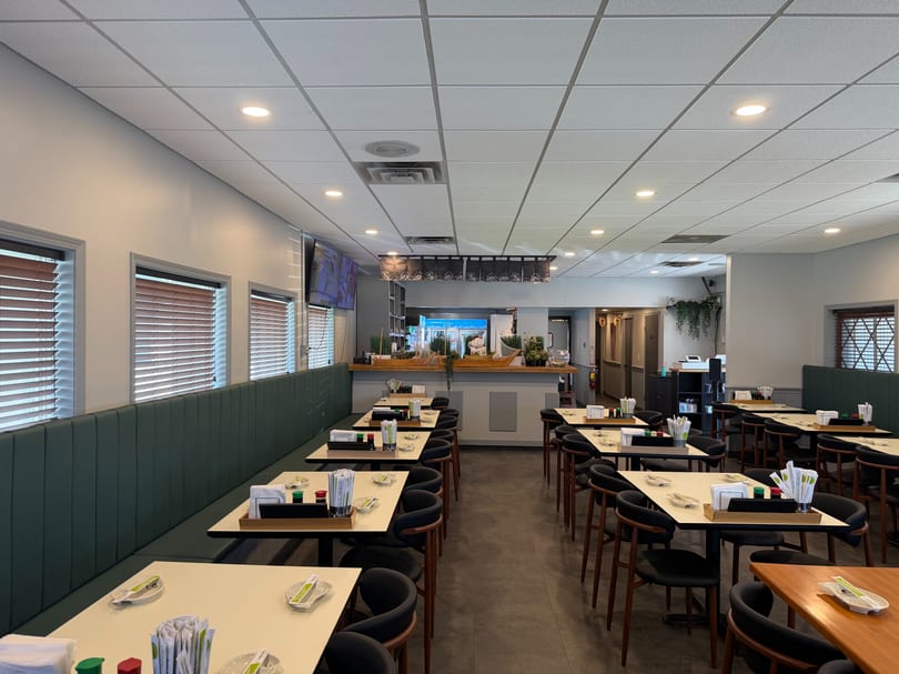 Empty restaurant dining room with green booth seating, light tables, and dark chairs, leading to a bar area.