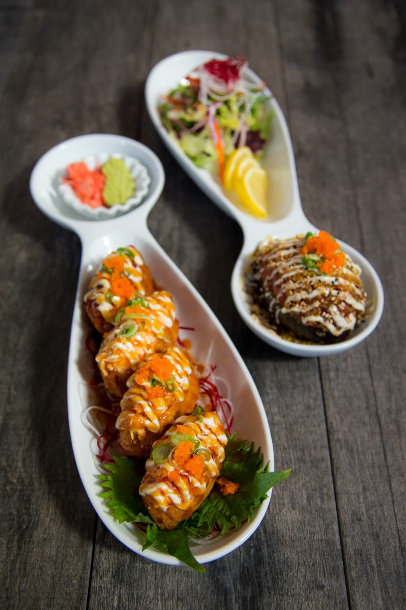 Three white dishes of Japanese appetizers, including fried rolls, salad, and a rice dish, on a wooden table.